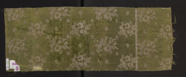 Fragment of a solid silk fabric, patterned with two joined branches bearing unfolded roses and small flowers arranged in an alternating rhythm. Fragment, 16 x 11.5 cm; overall cloth height 61 cm, width 23 cm. 2nd half of the 18th century, possibly Poland Grodno., A rectangular piece of fabric with a woven floral pattern lies flat against a dark background; the fabric shows a few areas of wear and repair and fraying along the edges. The textile is primarily olive green with lighter green and pale beige floral motifs repeated across it. The floral elements include stylized blossoms and leafy sprigs arranged in clusters and scattered small floral shapes. There is a vertical seam or fold near one end and a few irregular patches where the weave appears disturbed or mended. Loose threads and a small area of dense stitching are visible near one edge.