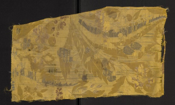 Fabric fragment, fragments of silk fabrics, patterned yellow silk damask, embroidered and brocaded with fanciful plant decoration. Damask substructure, satin with warp and grosgrain; decoration embellished with white weft and colored wefts. Elongated, frayed leaves form a background for smaller fanciful flowers brocaded with colored threads. Colors: white and yellow background. Embroidery with colored threads: sea green, light pink, dark pink, gold metallic thread. Edge width selvage: 0.3 cm; selvage made of thicker yellow threads. Early 18th century, Europe. Height 44 cm, width 25 cm., A rectangular piece of woven fabric with frayed edges lies against a dark background; the fabric is primarily golden yellow with patterns woven in a few shades including muted blue-gray and dull reddish-brown, showing a floral and leaf motif with overlapping large leaves and several clusters of rounded flower shapes, some areas of the weave appear worn or slightly abraded revealing horizontal thread var...