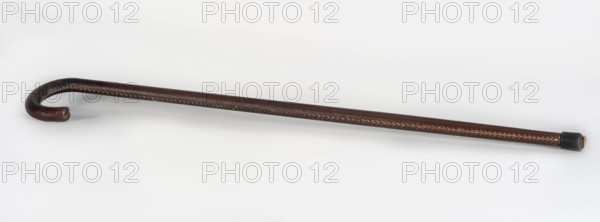 Walking stick, maker, wooden walking stick covered with brown leather along its entire length, rubber-coated foot; 20th century, Europe; men's fashion accessory; length 81 cm, width 7 cm., A single curved walking stick lies on a light background, showing a hooked handle and a straight shaft; the stick is primarily dark brown with lighter tan stitching or decorative inlay running along its length, the hooked end is rounded and the opposite tip is capped with a short, darker piece.