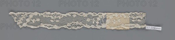 Lace fragment, lace with an ornament carried on two edges by a free tendril and carried diagonally through the central part, creating large fields filled with a net of large knitted meshes; mid-18th century, France; fabric; width 3.5 cm, length 28 cm., A long narrow strip of lace placed against a gray background, showing a delicate openwork pattern with a scalloped top edge and an irregular bottom edge; the lace features a honeycomb-like mesh interspersed with floral and leaf motifs and raised corded outlines, and a small patch of slightly darker fabric is attached near one end; visible colors include off-white and cream in the lace and patch, and gray in the background.