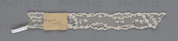 Lace fragment, lace with ornament carried on two edges with a free tendril, also carried diagonally through the central part so that large fields filled with a net with large knitted meshes are marked out; mid-18th century, France; fabric; width 3.5 cm, length 28 cm., A narrow strip of delicate lace in off-white stretches horizontally against a medium gray background, featuring a repeating floral and net pattern with scalloped edges; a small rectangular piece of light brown cardboard is attached near one end, partially covering the lace, and a short white ribbon tab protrudes from beneath the lace at the same end.