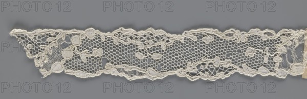 Lace fragment, mid-18th century, France. Lace with an ornament carried on two edges and a free tendril also carried diagonally through the central part, creating large fields filled with a net of large knitted meshes. Fabric; width 3.5 cm, length 28 cm., A long strip of delicate lace in off-white and cream tones stretches horizontally against a smooth gray background, featuring a hexagonal open net pattern interspersed with denser embroidered motifs resembling rounded shapes and small floral forms, with a scalloped uneven top and bottom edge, visible loose threads and slight fraying along some edges, and subtle variations in the off-white color where the thicker embroidered areas appear a touch darker than the finer netting.