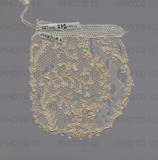 Lace fragment of a jabot, lace, 1st half of the 18th century, France; dimensions: length 10 cm, width 8 cm., A delicate piece of lace in a cream color hangs against a flat gray background, featuring an openwork net ground and an embroidered floral vine motif with leaves and small blossoms; the top edge shows a narrow band of tighter netting with a few attached strings, and the lace has an overall scalloped lower edge with curved outlines and varied density in the pattern.