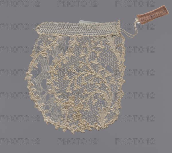 Lace — fragment of a jabot, maker, Kublicki Piottuch, Alfred 1861–1945 creator, lace, 1st half of the 18th century, France; material: lace; dimensions: length 10 cm, width 8 cm., A piece of delicate lace in a creamy off-white color with an open hexagonal mesh and embroidered floral and leafy motifs forming a rounded oval shape, a scalloped edge with small looped points, a straight top band with a tighter mesh, and a small fabric tag attached by thin cords in a muted brownish-pink color; the background is a uniform cool gray.