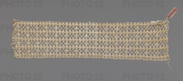 Fragment of crocheted lace — section of a belt, cream-colored, with motifs of semicircular shells and small rhombuses in rows. Lace fragment, width 7 cm, length 31 cm. 1st half of the 19th century, Germany ., A rectangular piece of openwork fabric with a repeating diamond-like lattice pattern in a pale beige color laid flat against a smooth medium gray background, with uneven edges and a loose thread extending from one corner, and a small rectangular tag attached near one corner in a light brown tone.
