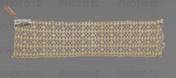 Fragment of crocheted lace — section of a belt, cream-colored, with motifs of semicircular shells and small rhombuses in rows. Lace fragment, width 7 cm, length 31 cm. 1st half of the 19th century, Germany ., A rectangular piece of openwork fabric with a netlike pattern made from a beige yarn stretches horizontally against a solid gray background, showing a repeating diamond-shaped lattice with textured clusters at the intersections and some loose threads at the edges; a small fabric tag and a narrow leather or tan-colored tag are attached at the upper left corner.