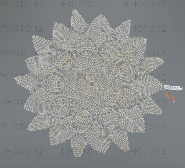 Napkin, napkin, lace napkin, white, round with large triangular teeth along the edge, concentric and radial geometric ornament, 20th century, Poland, fabric, lace, diameter 33 cm, A round crocheted doily with a lacy floral pattern in off-white and pale cream tones laid flat against a solid medium-gray background, featuring a central circular motif surrounded by concentric rings of openwork and petal-like shapes, with a scalloped outer edge of pointed leaf motifs; a small fabric tag and a small rectangular tag in brown are attached near the edge.