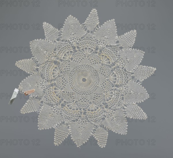 Napkin, napkin, lace, lace napkin, white, round with large triangular teeth along the edge; concentric and radial geometric ornament; 20th century, Poland; fabric, lace; diameter 33 cm, A circular, lace-like doily with an intricate radial pattern forming petal-shaped points around the edge, displayed flat against a smooth gray background; the doily is primarily off-white with subtle hints of pale yellow woven into its center and some inner rings, and there is a small clothespin-like clip attached near the left edge that is light gray with a touch of reddish-brown material nearby.