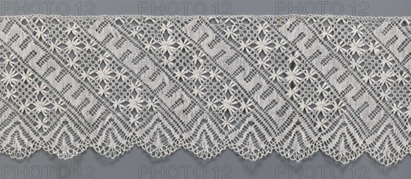 Lace fragment, bobbin lace. White, section of a strip with a pattern of diagonal stripes with rosettes and a meander; one edge serrated. 19th century, Europe. Width 17.5 cm, length 395 cm., A length of lace with an open, geometric floral pattern lies against a smooth background. The lace features repeating diagonal bands of mesh and solid woven sections forming stylized shapes, interspersed with daisy-like floral motifs with petal clusters. The lower edge of the lace has a scalloped border with triangular, fan-like motifs and delicate looped trim. Visible colors include off-white or cream for the lace and a medium gray background.