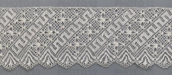 Lace fragment, bobbin lace; section of a strip. White, with a pattern of diagonal stripes with rosettes and a meander. One edge is serrated. 19th century, Europe. Width 17.5 cm, length 395 cm., A band of lace in off-white stretches across a gray background, featuring a repeating geometric pattern of zigzag and diamond shapes interspersed with floral motifs; the lace has openwork areas with fine net-like stitching and solid woven sections, and the lower edge forms a scalloped outline with triangular arches and delicate picot-like loops.