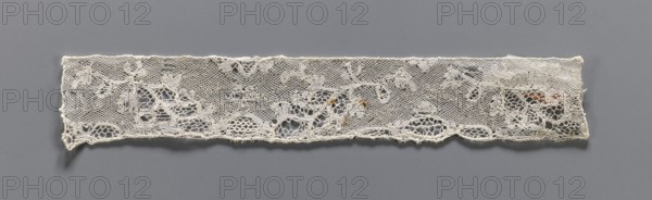 Lace fragment, bobbin lace Valenciennes, white, continuous floral pattern with Rococo motifs, 18th century, width 4 cm, length 23 cm, A horizontal strip of delicate lace with a floral and leaf pattern laid against a smooth background; the lace is off-white with subtle variations in tone and a few small areas of brownish discoloration, showing open mesh sections and more densely stitched floral motifs, scalloped lower edge, and irregular frayed ends, all set on a flat medium gray backdrop.