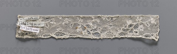 Lace fragment, bobbin lace, Valenciennes lace. Fragment of white bobbin lace with a continuous floral pattern and Rococo motifs. 18th century. Width 4 cm, length 23 cm., A narrow strip of lace with a floral and netted pattern lies horizontally against a solid background, showing a delicate open weave with floral motifs and scalloped lower edge; the lace appears off-white to cream with a few small darker discolorations, and the background is a uniform gray.