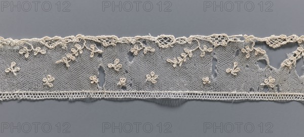 Fragment of needle lace of the point d'Alençon type. Cream needle lace with a mesh background of twigs, leaves, stars, and rhombuses. 18th century, possibly France. Width 4.7 cm, length 73.5 cm., provenance includes Henryków Heinrichau., A narrow strip of delicate lace in off-white and cream tones stretches horizontally against a smooth grey background, featuring a fine hexagonal net ground with a scalloped upper edge and a straight lower edge, decorated with a scattering of small floral motifs and leaf-like loops made of thicker corded thread, showing a few irregular holes and worn areas in the net and some loose thread ends, with subtle shadows beneath the lace creating a slight sense of depth.