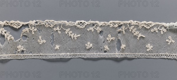 Fragment of needle lace of the point d'Alençon type. Cream, with a mesh background of twigs, leaves, stars, rhombuses. 18th century, France . Needle lace, width 4.7 cm, length 73.5 cm., Repossession provenance noted; Henryków German: Heinrichau mentioned., A strip of delicate openwork lace in off-white and cream tones lies horizontally against a smooth gray background; the lace features a fine hexagonal net ground with a scalloped top edge and a straight bottom edge, decorated with a scattering of small raised floral motifs and clusters of looped thread creating tiny flowers and leaves, along with a few irregular holes and worn areas; the top border shows a slightly more textured, knotted lace pattern while the bottom border has a narrow band of denser looping.