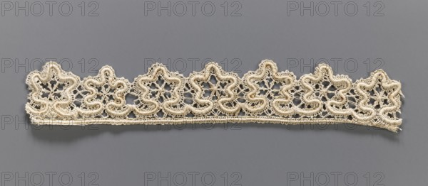 Lace fragment, needle-stitched lace, upper edge straight, lower edge serrated to match the motifs. The pattern of the motifs and the upper edge are emphasized by thickly sewn threads. 19th century, Europe. Height 4 cm, width 27 cm., A horizontal strip of delicate lace with a repeating floral-like scalloped pattern rests against a solid background. The lace is off-white or cream colored with a slightly darker beige tone in some areas, showing a textured, looped cord outlining each petal-like shape and finer openwork filling the interior. The background is a smooth grey, and the lace casts a soft shadow beneath it. The strip ends are slightly frayed, and the overall appearance shows fine threadwork and small interconnected loops forming a lace border.