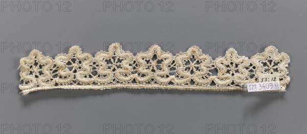 Lace fragment, needle-stitched lace, upper edge straight, lower edge serrated to match the motifs. The pattern of the motifs and the upper edge are emphasized by thickly sewn threads. 19th century, Europe. Height 4 cm, width 27 cm., A narrow length of intricately patterned lace in an off-white or cream color is shown laid horizontally against a flat gray background; the lace has a scalloped top edge with a repeating floral-like motif formed by openwork loops and dense embroidered areas, and a straight lower edge with a fine woven border; there are small areas of slightly darker discoloration within the lace and a small paper tag attached near one end that contrasts with white and light tones.
