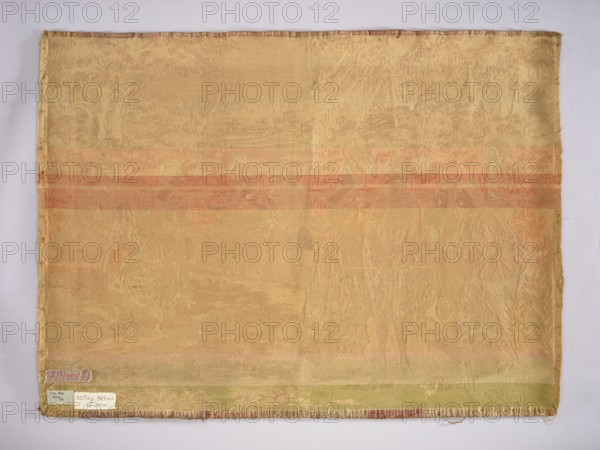 Jacquard tapestry by Mariano Alonso Pérez 1857–1930, co-created by an unknown manufacturer. Depicts ladies and gentlemen in Louis XVI–era costumes watching a lady jumping rope, set in a park landscape. No border. Monochromatic colors in shades of beige and ochre. 19th/20th century, Europe. Materials: fabrics; height 73 cm, width 101 cm., A rectangular woven textile laid flat against a light background, showing a faded overall yellow-beige color with horizontal bands of muted orange and pale green across its width; the fabric surface has a subtle raised pattern depicting indistinct figures and foliage in slightly lighter and darker shades of the base color, with fringed short edges and stitched borders visible along the sides; a few small paper labels are attached near one corner showing contrasting pale and darker tones.