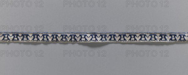 Gallon, galloons haberdashery, origin unknown, plant ornaments, 19th century, Europe, fabric, haberdashery, width 1.3 cm, length 49.5 cm, A horizontal woven band runs across the image against a smooth gray background, showing a repeating geometric motif in dark blue on an off-white base with thin dark blue borders along its top and bottom edges; the band has a slightly textured, fibrous appearance and casts a soft shadow on the background, and the visible colors are dark blue, off-white, and gray.