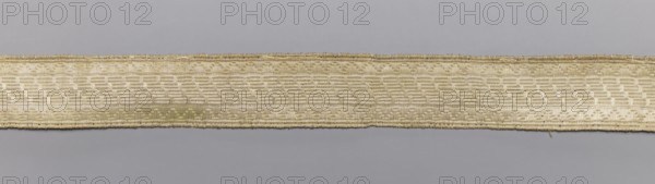 Gallon, galloons haberdashery, geometric ornaments, white warp, weft - wire - drawn thread. Faint ornament, broken line at the edge, geometric ornament in the center: warp stripes interrupted by diagonal, stepped lines. 19th century, Europe. Fabric haberdashery, width 2.2 cm, length 77 cm., A horizontal woven band runs across the image against a plain background, showing a textured pattern of interlaced threads and a slightly raised border along its top and bottom edges; the band features a repeating rectangular weave and some irregularities in thread thickness, with loose fiber ends visible at one point, and the background appears smooth and even. The visible colors are beige, cream, light tan, and gray.