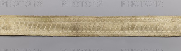 Gallon, galloons haberdashery, geometric ornaments, white warp, weft - wire - drawn thread. Faint ornament, broken line at the edge, geometric ornament in the center: warp stripes interrupted by diagonal, stepped lines. 19th century, Europe. Fabric, haberdashery, width 2.2 cm, length 77 cm., A long woven ribbon with a textured pattern runs horizontally across the image against a smooth background; the ribbon shows a repeating basketweave-like design with a slightly raised border along the top and bottom edges, and a few small irregularities in the weave. The ribbon is primarily beige and cream with subtle variations of light tan and off-white creating a faint sheen, and the background is a uniform gray.