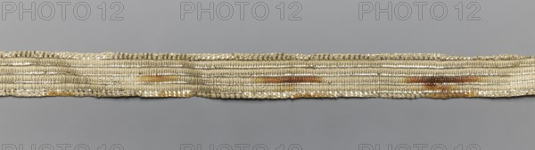 Gallon, haberdashery, origin unknown, geometric ornaments, 19th century, Poland, fabric, width 1.5 cm, length 71 cm, A long narrow woven band laid horizontally against a gray background, showing a textured pattern of tightly braided and looped threads; the band appears primarily off-white or beige with a metallic sheen, and has areas of muted gold and brownish-orange discoloration along its length, with subtle highlights of silver from reflective threads and faint shadows that emphasize the ridged, ribbed surface.