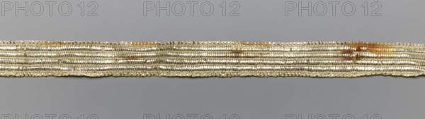 Gallon, gallons haberdashery, origin unknown, geometric ornaments, 19th century, Poland, fabric, haberdashery, width 1.5 cm, length 71 cm, A narrow horizontal strip of metallic braided material stretches across a plain background, showing a woven texture with a repeating ribbed pattern and some irregularities along its edges; the strip appears predominantly silvery with areas of pale gold and a few rust-colored brown spots, and the background is a uniform gray.