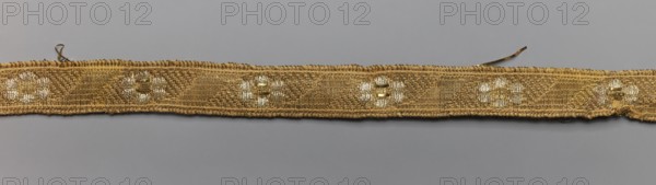 Gallon, gallons haberdashery, origin unknown, plant ornaments, 18th century, Poland, Drzewica, fabric haberdashery, width 1.4 cm, length 40 cm, A narrow woven strip lies horizontally across a neutral background, showing a textured braided surface with a repeating floral-like motif; the strip is primarily gold with accents of lighter metallic tones and small areas of white sheen, and it has slightly irregular edges with a few loose thread ends visible.