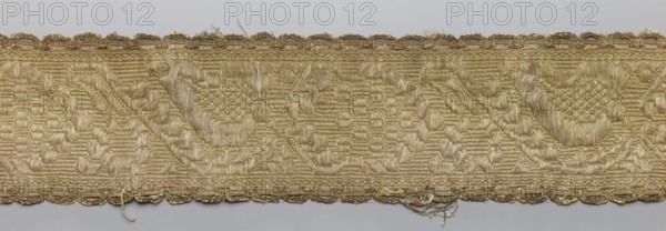 Gallon, gallons haberdashery, origin unknown, plant ornaments, 19th century, Lithuania, fabric, haberdashery, width 9 cm, length 57 cm, A horizontal strip of woven fabric with a repeating geometric pattern in shades of gold and beige, showing a raised, textured weave and subtle sheen; the top and bottom edges have a scalloped decorative border in metallic gold threads with some loose fibers and fraying; the central field displays repeating triangular and circular motifs created by variations in weave height and direction, giving a mix of matte and shiny surfaces in gold, beige, and light tan tones.