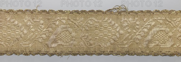 Gallon, haberdashery, origin unknown, plant ornaments, 19th century, Lithuania, fabric, width 9 cm, length 57 cm, A narrow woven trim with a repeating decorative pattern in shades of gold and beige, showing a textured background of horizontal ribbing and small geometric motifs, framed by a scalloped edging that has loose threads and slight fraying; the weave includes raised areas forming arc-like floral shapes and clustered textured centers, with subtle variations in sheen where light reflects off the silky threads, and a pale gray background visible above and below the trim.