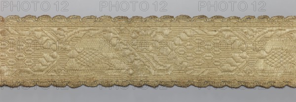 Gallon, haberdashery, origin unknown, 19th century, possibly Lithuania, fabric, haberdashery, width 8.5 cm, length 60 cm, A horizontal strip of woven fabric in shades of beige and light gold with a repeating ornate pattern and a scalloped edge; the weave shows textured motifs including curved and geometric shapes with areas of shinier and more matte threads, and the background behind the strip is a smooth pale gray.