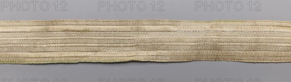 Gallon, haberdashery trim, geometric ornaments, 19th century, Europe, fabric, width 3 cm, length 54 cm, purchase noted provenance., A long narrow woven textile strip lays horizontally across a smooth background, showing a tightly woven pattern of parallel horizontal ribs and subtle raised ridges; the fabric appears slightly uneven in places with gentle waves along its edges and a finished hem running the length. The visible colors include shades of beige and cream throughout the textile, with hints of light green along the lower edge and tiny touches of pale brown in the woven threads, set against a cool gray background.
