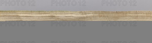 Gallon, haberdashery; geometric ornaments; 19th century, Europe; fabric; width 3 cm, length 54 cm; used as a haberdashery trim., A horizontal scene dominated by a long, narrow woven band with visible texture and layered ridges, showing natural beige and tan fibers with subtle greenish and light brown accents along the upper edge, set against a smooth pale gray background above and a solid mid-gray area below.