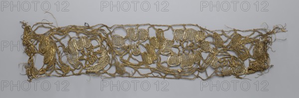 Lace fragment, lace, 2nd half of the 18th century, possibly Flanders, fabric, width 7.5 cm, length 35 cm., A long, narrow, irregularly shaped openwork piece made of metallic thread in gold tones laid flat against a smooth light gray background, with tangled loops and braided strands forming a loose network and a few denser woven patches, frayed loose ends and wispy threads extending from the edges, and subtle variations in the gold appearance where the threads catch the light.