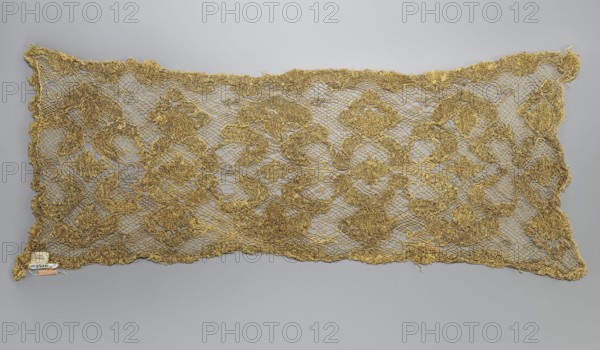 Fragment of lace, lace, 17th/18th century, Belgium, fabric, lace, width 31 cm, length 76 cm, A rectangular piece of openwork fabric laid flat against a neutral background, showing a repeating ornate floral and scroll-like pattern made from metallic threads in shades of gold and bronze, with the ground formed by a net-like mesh of thin threads and the decorative motifs appearing raised and textured; the edges are irregular and scalloped, and a small label-like object is attached near one corner.