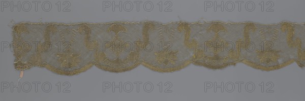 Lace fragment attributed to Kublicki Piottuch or Alfred 1861–1945. Lace strip finished with flat, arched teeth on one side and straight on the other. In the divisions corresponding to the teeth, an alternating flower calyx and a rhombus motif are placed, separated by parenthetical geometric forms. 18th century, France. Material: fabric, lace; width 17.5 cm, length 308 cm., A narrow strip of lace with a scalloped lower edge and a straight upper edge, featuring a repeating embroidered pattern of ornamental shapes and floral motifs; the lace is primarily gold and appears set against a gray background, with areas of fine netting and denser embroidered sections forming curved S-shaped scrolls, diamond-like lattice elements, and stylized flower clusters; the gold thread shows slight variations in sheen and texture, and there are a few loose threads and frayed edges visible along the top and at one end.
