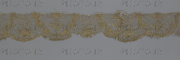 Lace fragment, lace strip finished with flat arched teeth on one side and straight on the other. In the divisions corresponding to the teeth, an alternating flower calyx and a rhombus motif are placed, separated by parenthetical geometric forms. 18th century, France; lace, fabric; width 17.5 cm, length 308 cm., A narrow strip of lace-like fabric with a repeating ornamental pattern rests against a plain background; the fabric is primarily golden with variations of muted gold and tan, and the background is a cool gray. The lace shows a mesh ground with a scalloped lower edge and an upper straight edge, and the decorative motif alternates between curving scroll shapes and floral clusters connected by small geometric lattice elements. The scroll motifs are rounded and sinuous, the floral motifs have layered petal forms, and there are areas where threads are frayed or missing, creating small gaps. The overall texture appears delicate and airy with denser embroidered outlines and open netted...