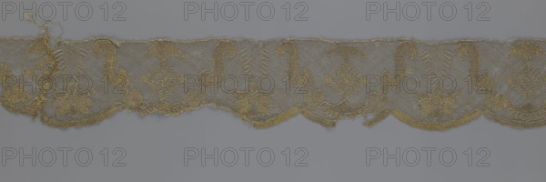 Lace fragment, lace strip finished with flat arched teeth on one side and straight on the other; in the divisions corresponding to the teeth an alternating flower calyx and a rhombus motif are placed, separated by parenthetical geometric forms. 18th century, France; fabric; width 17.5 cm, length 308 cm., A strip of delicate lace with a repeating decorative pattern rests against a plain background; the lace shows floral motifs and curved ornamental shapes connected by a net-like mesh, with scalloped edges along one side and some loose threads and fraying at irregular points; visible colors include shades of gold and beige in the lace set against a muted gray background.