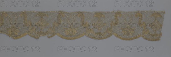 Lace fragment, lace strip finished with flat arched teeth on one side and straight on the other; in the divisions corresponding to the teeth, an alternating flower calyx and a rhombus motif are placed, separated by parenthetical geometric forms. 18th century, France. Materials: lace, fabric. Dimensions: width 17.5 cm, length 308 cm., A horizontal strip of delicate lace with scalloped lower edges and a repeating ornamental pattern featuring curved S-shaped motifs, floral forms, and diamond-like lattice medallions, all rendered in a metallic golden thread against a muted gray background; the lace shows areas of open netting and denser embroidered sections, with fine tonal variations of gold and subtle hints of darker and lighter gray where the background shows through.