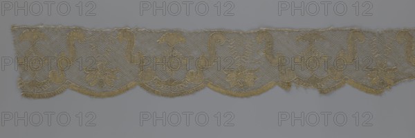 Lace fragment, attributed to Kublicki Piottuch, Alfred 1861–1945; lace strip finished with flat, arched teeth on one side and straight on the other. In the divisions corresponding to the teeth, an alternating flower calyx and a rhombus motif are placed, separated by parenthetical geometric forms. 18th century, France; material: lace, fabric; width 17.5 cm, length 308 cm., A narrow strip of delicate lace in shades of beige and light gold runs horizontally across the image, featuring a repeating scalloped lower edge and a fine netted background; within the netting are a few ornate embroidered motifs including curved scroll shapes, floral forms with petal-like elements, and diamond-like decorative panels, all rendered in the same beige-gold thread; the lace shows slight irregularities along its top edge and a few loose threads, set against a smooth muted gray-blue background.