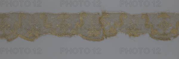 Lace fragment, lace strip finished with flat arched teeth on one side and straight on the other; in the divisions corresponding to the teeth an alternating flower calyx and a rhombus motif are placed, separated by parenthetical geometric forms. 18th century, France. Materials: lace, fabric. Dimensions: width 17.5 cm, length 308 cm., A horizontal strip of delicate lace with a floral and scrolling pattern lies against a flat background; the lace shows a repeating design of flower motifs and sinuous scrolls connected by a fine net ground, with scalloped lower edges and some frayed threads and worn areas; visible colors include shades of muted gold and beige in the lace and a cool gray background.