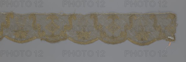 Lace fragment by Kublicki Piottuch, Alfred 1861–1945; lace strip finished with flat, arched teeth on one side and straight on the other. In the divisions corresponding to the teeth, an alternating flower calyx and a rhombus motif are placed, separated by parenthetical geometric forms. 18th century, France; lace fabric; width 17.5 cm, length 308 cm., A horizontal strip of lace with a scalloped lower edge featuring repeating ornate motifs; the lace has a mesh background with embroidered curving S-shaped forms, umbrella-like shapes, floral designs, and decorative zigzag elements. The embroidery thread appears gold against a gray background, and the piece shows some frayed threads along the top edge and scallops. There is a small paper tag and a short string attached near one end; the overall colors visible are gold and gray.