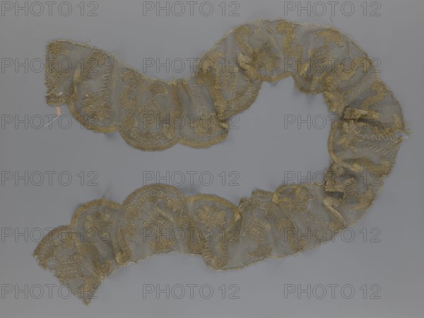 Lace fragment, lace strip finished with flat, arched teeth on one side and straight on the other. In the divisions corresponding to the teeth, an alternating flower calyx and a rhombus motif are placed, separated by parenthetical geometric forms. 18th century, France. Materials: lace, fabric. Dimensions: width 17.5 cm, length 308 cm., A sinuous length of lace arranged in a loose S-shape on a gray background, the lace appearing delicate and semi-transparent with scalloped edges and a repeating floral and scroll motif; the lace is primarily gold in color with variations in tone where threads cluster, and the background is a uniform muted gray.