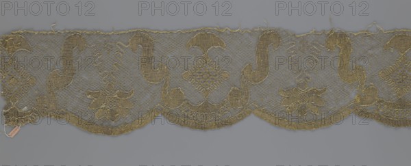 Lace fragment by Kublicki Piottuch, Alfred 1861–1945. The lace strip is finished with flat, arched teeth on one side and straight on the other. In the divisions corresponding to the teeth, an alternating flower calyx and a rhombus motif are placed, separated by parenthetical geometric forms. 18th century, France. Materials: lace, fabric. Width 17.5 cm; length 308 cm., A horizontal strip of ornate lace with a scalloped lower edge displayed against a plain background, showing a repeating embroidered pattern of curving paisley-like shapes, diamond motifs, and floral clusters; the lace has an open netted ground with denser, textured embroidery forming the decorative elements and a slightly frayed top edge with loose threads; visible colors include muted gold thread for the embroidery and a cool gray background.