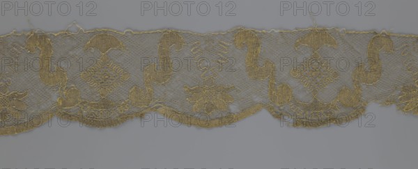Lace fragment, lace strip finished with flat arched teeth on one side and straight on the other. In the divisions corresponding to the teeth, an alternating flower calyx and a rhombus motif are placed, separated by parenthetical geometric forms. 18th century, France. Materials: lace, fabric. Dimensions: width 17.5 cm, length 308 cm., A horizontal strip of decorative textile with an open net-like background in gray and a repeating embroidered pattern in golden-yellow; the embroidered motifs include sweeping curved shapes, umbrella-like forms, diamond-shaped lattice elements, floral or leaf clusters, and scalloped lower edges with fringe. The net background appears slightly translucent and gray, while the embroidery thread is a metallic golden-yellow that outlines and fills the motifs, creating contrast between the two colors. The textile shows some loose threads and small irregularities along the top and bottom edges, and the overall composition runs horizontally across a neutral gray b...