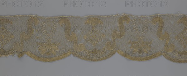 Lace fragment, maker unknown possibly Kublicki Piottuch, Alfred, 1861–1945 noted, lace strip finished with flat arched teeth on one side and straight on the other; divisions corresponding to the teeth feature alternating flower calyx and rhombus motifs separated by parenthetical geometric forms. 18th century, France; lace, fabric; width 17.5 cm, length 308 cm., A horizontal strip of lace with a scalloped lower edge displaying a repeating ornate pattern in gold and beige tones against a gray background; the lace features a mesh netting base with embroidered curved motifs, floral shapes, and diamond-like medallions, along with decorative looping stitches and a textured trimmed border, showing variations of gold, beige, gray, and hints of brown.