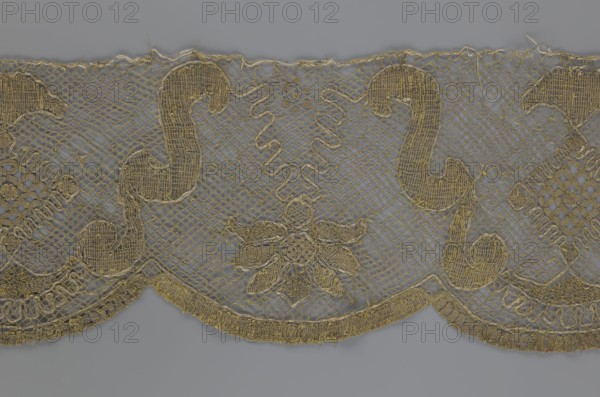 Lace fragment;, The lace strip is finished with flat, arched teeth on one side and straight on the other. In the divisions corresponding to the teeth, an alternating flower calyx and a rhombus motif are placed, separated by parenthetical geometric forms. 18th century, France. Materials: lace, fabric. Dimensions: width 17.5 cm, length 308 cm., The image shows a decorative textile strip with an open net-like background in gray and gold tones, featuring ornate gold embroidered motifs including curved scroll shapes and a central floral element; the lower edge has a scalloped border with dense gold embroidery, and there are a few small loose threads and irregularities along the top edge.