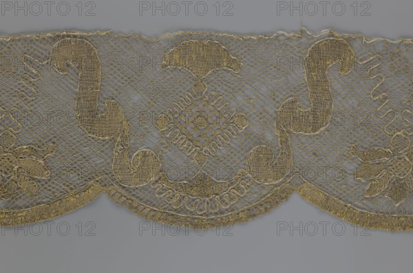 Lace fragment, lace. The lace strip is finished with flat, arched teeth on one side and straight on the other. In the divisions corresponding to the teeth, an alternating flower calyx and a rhombus motif are placed, separated by parenthetical geometric forms. 18th century, France. Fabric, width 17.5 cm, length 308 cm., A delicate piece of lace with a scalloped lower edge shown against a gray background, featuring a repeating ornamental pattern of curving paisley-like shapes and a central diamond-shaped motif, all rendered in a golden-beige thread on a fine open net ground; several floral and leafy elements are integrated into the design, and the embroidery displays variations in texture with denser stitches in the motifs and finer mesh in the surrounding areas, the colors visible being gold, beige, and gray.