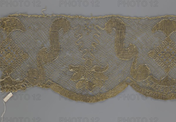 Lace fragment, lace strip finished with flat arched teeth on one side and straight on the other; in the divisions corresponding to the teeth an alternating flower calyx and a rhombus motif are placed, separated by parenthetical geometric forms. 18th century, France; fabric; width 17.5 cm, length 308 cm., A length of patterned lace fabric laid flat showing a scalloped lower edge and an upper straight edge, with an open net ground and larger embroidered motifs integrated into the mesh. The lace features a repeating design of curved scroll-like shapes and floral-inspired clusters, with denser textured stitching outlining the motifs and small decorative openwork areas at intervals. The fabric appears delicate with some loose threads and frayed edges visible along the top and bottom. The visible colors in the image are gold tones for the lace against a neutral gray background, with slight variations in the gold where stitching density changes.