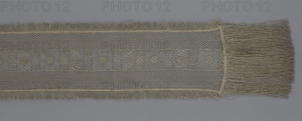 A so-called Saxon lace belt, lace, belts, clothing accessories, men's clothing, fashion, provenance unknown. The central part is a continuous belt with a geometric ornament; among the motifs there is a circle containing an hourglass. The side belts of similar width are permanently connected. The entire belt is trimmed with fringe. 17th/18th century, Poland, lace, clothing accessories, height 270 cm, width 25 cm., A horizontal strip of woven fabric with fringed edges running along the top and bottom and a longer fringe at the right end, the textile shows a central patterned band with an open lattice structure flanked by areas of tighter net-like weave; the material appears in shades of beige and gray with variations of light tan and muted brown in the threads, set against a smooth gray background.