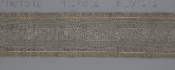 A so-called Saxon lace belt, creator, lace belt for men's clothing; central continuous belt with a geometric ornament including a circle containing an hourglass; side belts of similar width permanently connected; entire belt trimmed with fringe; 17th–18th century, Poland; lace clothing accessory; height 270 cm, width 25 cm., A long horizontal strip of woven fabric with fringed edges, showing a repeated decorative pattern along its center; the piece displays a textured mesh background with a band of geometric and circular motifs, and fringes at the top and bottom. The visible colors are shades of gray, muted beige, and pale gold.